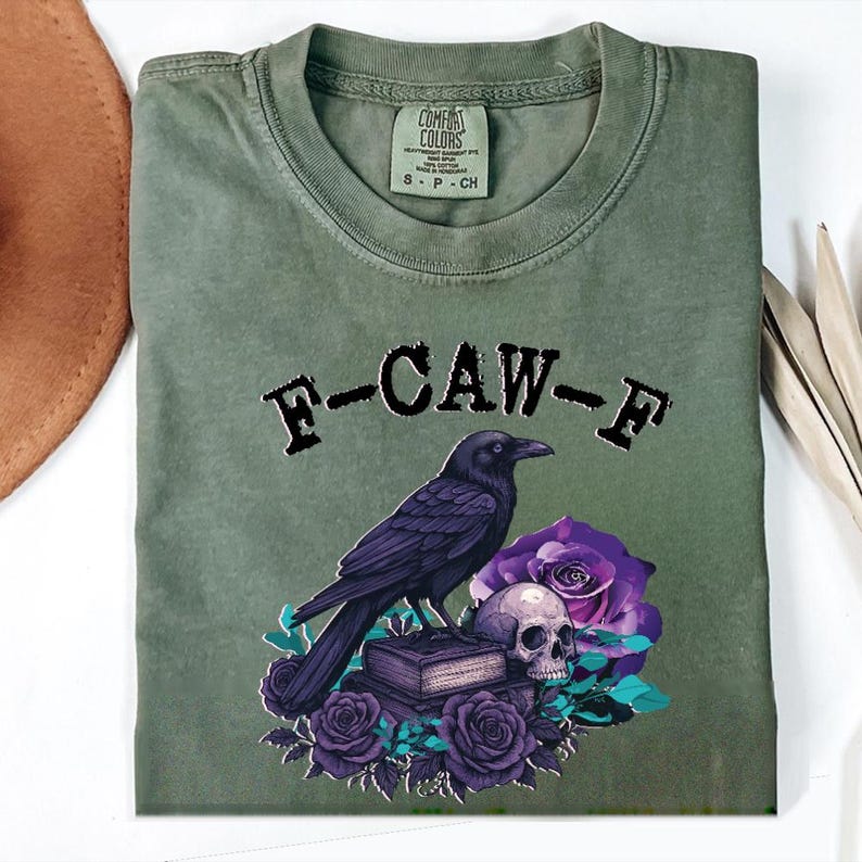May include: Olive green t-shirt with a graphic featuring a black crow perched above a skull, purple roses, and the text "F-CAW-F". The shirt is folded, showing the Comfort Colors label.