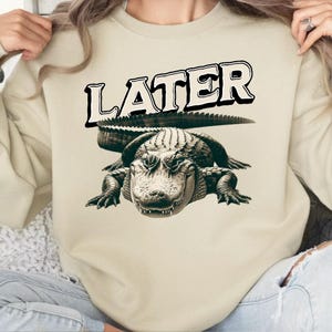 May include: A beige sweatshirt with the word "LATER" printed in black on the front. A black and white alligator graphic is below the text.