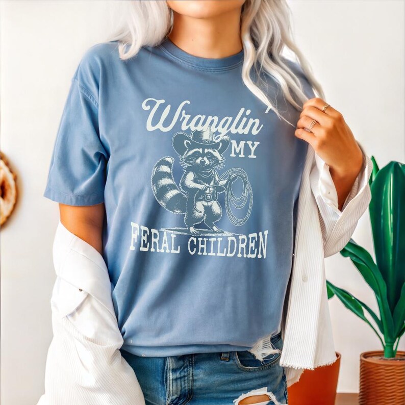 May include: A light blue t-shirt with a graphic of a raccoon wearing a cowboy hat and holding a lasso. The text "Wranglin' My Feral Children" is printed on the shirt.