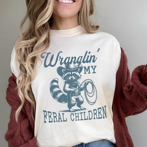 May include: A white t-shirt with a blue graphic of a raccoon wearing a cowboy hat and holding a lasso. The text "Wranglin' My Feral Children" is printed on the shirt.