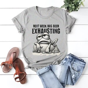 May include: A gray t-shirt with a cartoon frog and the text "Next week has been exhausting".