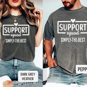 May include: Two dark grey t-shirts with the text "SUPPORT squad SIMPLY-THE-BEST" in a white rectangular box. One shirt is labeled "DARK GREY HEATHER" and the other is labeled "PEPPER". The shirts are worn by two people.