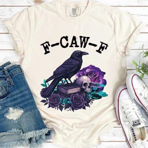 May include: Cream-colored t-shirt featuring a graphic of a black raven perched on a stack of books, a skull, and purple roses. The text "F-CAW-F" is printed above the graphic. The shirt is paired with denim shorts and white sneakers.