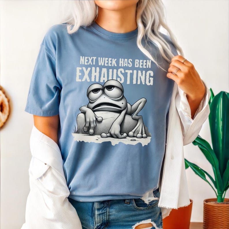May include: A light blue t-shirt with a cartoon frog wearing a tired expression. The shirt has the text "Next week has been exhausting" printed on it.