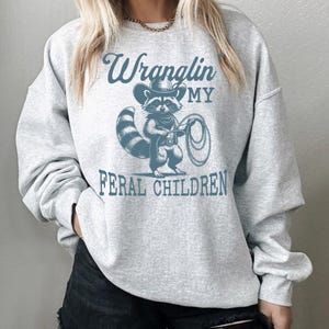 May include: Gray sweatshirt with a blue graphic of a raccoon wearing a cowboy hat and holding a lasso. The text "Wranglin' My Feral Children" is printed below the raccoon.
