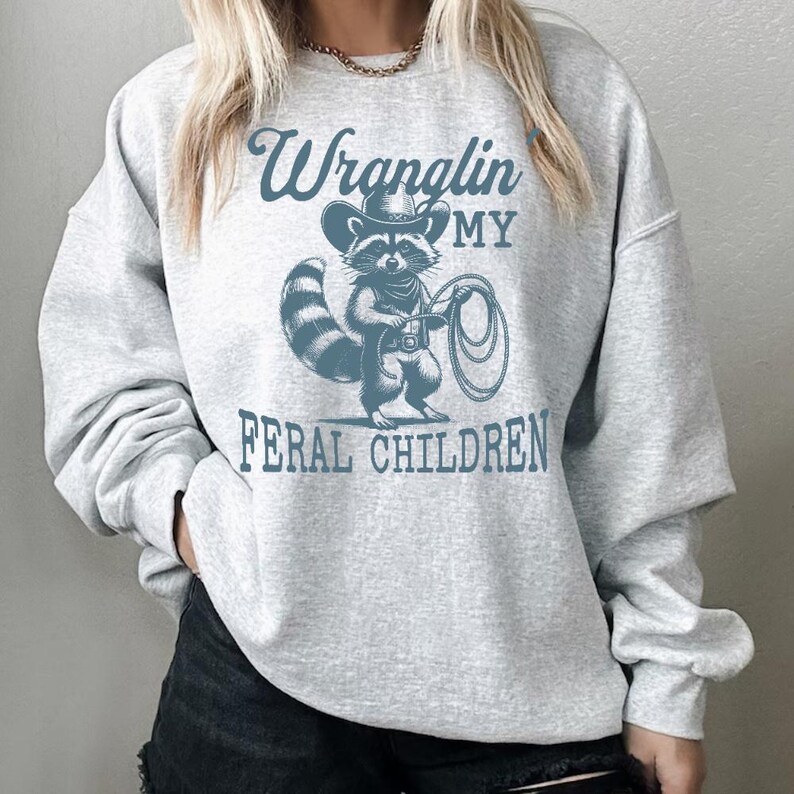 May include: Gray sweatshirt with a blue graphic of a raccoon wearing a cowboy hat and holding a lasso. The text "Wranglin' My Feral Children" is printed below the raccoon.