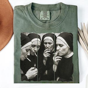 May include: Olive green Comfort Colors t-shirt featuring a black and white graphic of four nuns smoking cigarettes. The shirt has a crew neck and short sleeves. The Comfort Colors tag is visible at the neckline.