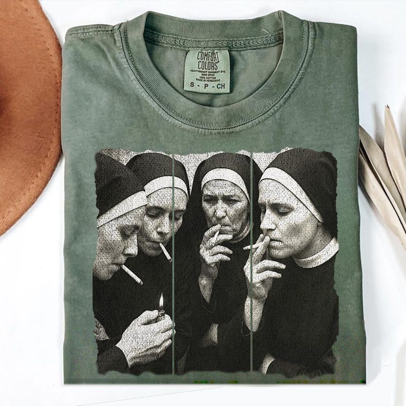 May include: Olive green Comfort Colors t-shirt featuring a black and white graphic of four nuns smoking cigarettes. The shirt has a crew neck and short sleeves. The Comfort Colors tag is visible at the neckline.