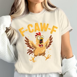 May include: Cream-colored t-shirt featuring a cartoon chicken with open wings and a surprised expression. The text "F-CAW-F" is printed above the chicken in yellow. The chicken has brown wings, a red comb, and yellow feet.