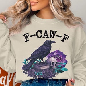 May include: Cream-colored sweatshirt with a graphic featuring a black crow, skull, purple roses, and the text "F-CAW-F". The design has a gothic or Halloween theme.