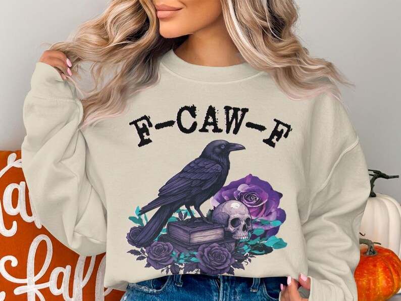 May include: Cream-colored sweatshirt with a graphic featuring a black crow, skull, purple roses, and the text "F-CAW-F". The design has a gothic or Halloween theme.