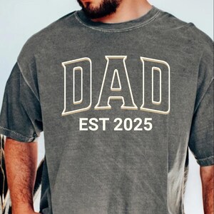May include: Dark gray t-shirt with the word "DAD" in a white and tan arched font. Below the word "DAD" is the text "EST 2025" in white. The shirt has short sleeves and a crew neck.
