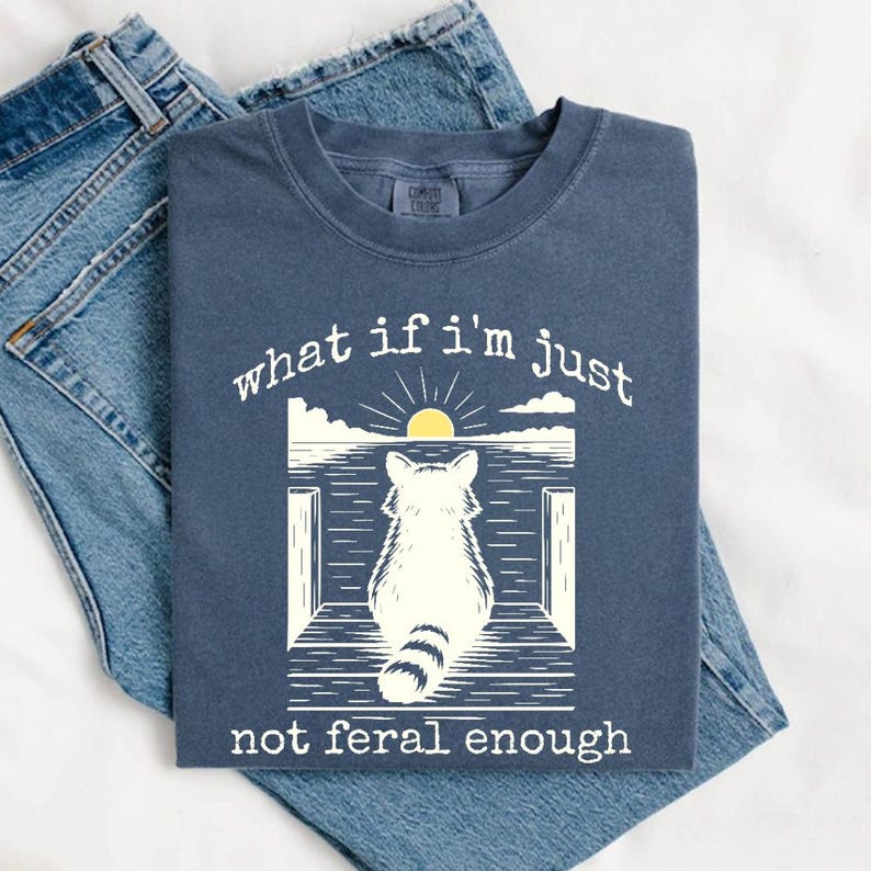 May include: A navy blue t-shirt with a white graphic of a raccoon sitting on a pier with the text "what if i'm just not feral enough".