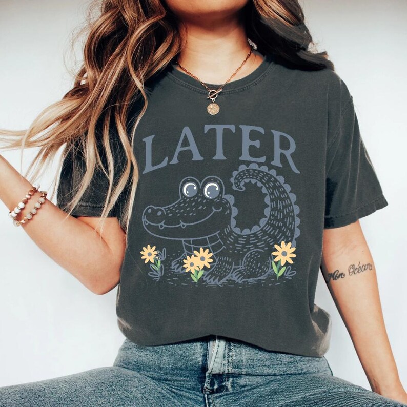 May include: A dark grey t-shirt featuring a cartoon alligator and the word "LATER" printed on it. The alligator is green and has large eyes. It is surrounded by yellow flowers.