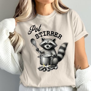 May include: A beige t-shirt with a black and white illustration of a raccoon stirring a pot over a campfire. The text "Pot Stirrer" is printed above the raccoon.