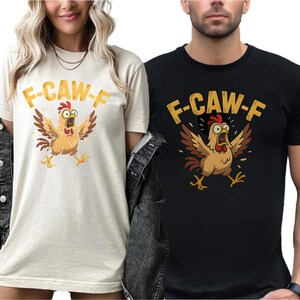 May include: Two t-shirts, one cream and one black, each featuring a cartoon chicken with outstretched wings and the text "F-CAW-F" in gold. The chicken is brown, with a red comb and yellow feet.
