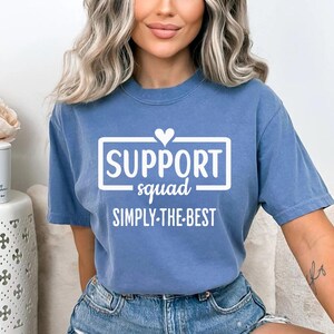 May include: A blue t-shirt with the words "SUPPORT squad SIMPLY THE BEST" in white. The text is inside a white rectangle with a heart above the word "SUPPORT". The shirt has short sleeves and a crew neck.