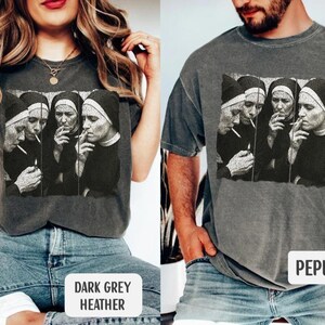 May include: Dark grey heather and pepper-colored t-shirts featuring a black and white graphic of four nuns smoking cigarettes. The image is printed on the front of the shirts. The shirts are displayed on two people.