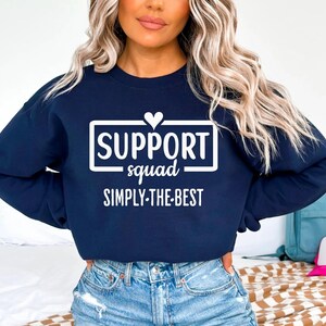 May include: Navy blue sweatshirt with the text "SUPPORT squad SIMPLY-THE-BEST" in white. The word "SUPPORT" is in a white rectangle with a small white heart above it. The sweatshirt is a crew neck style.