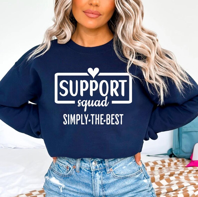 May include: Navy blue sweatshirt with the text "SUPPORT squad SIMPLY-THE-BEST" in white. The word "SUPPORT" is in a white rectangle with a small white heart above it. The sweatshirt is a crew neck style.