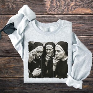 May include: Light gray sweatshirt featuring a black and white graphic of four nuns smoking cigarettes. The image is a stylized, artistic representation. The sweatshirt is folded on a wooden surface, with a pair of black sunglasses visible.