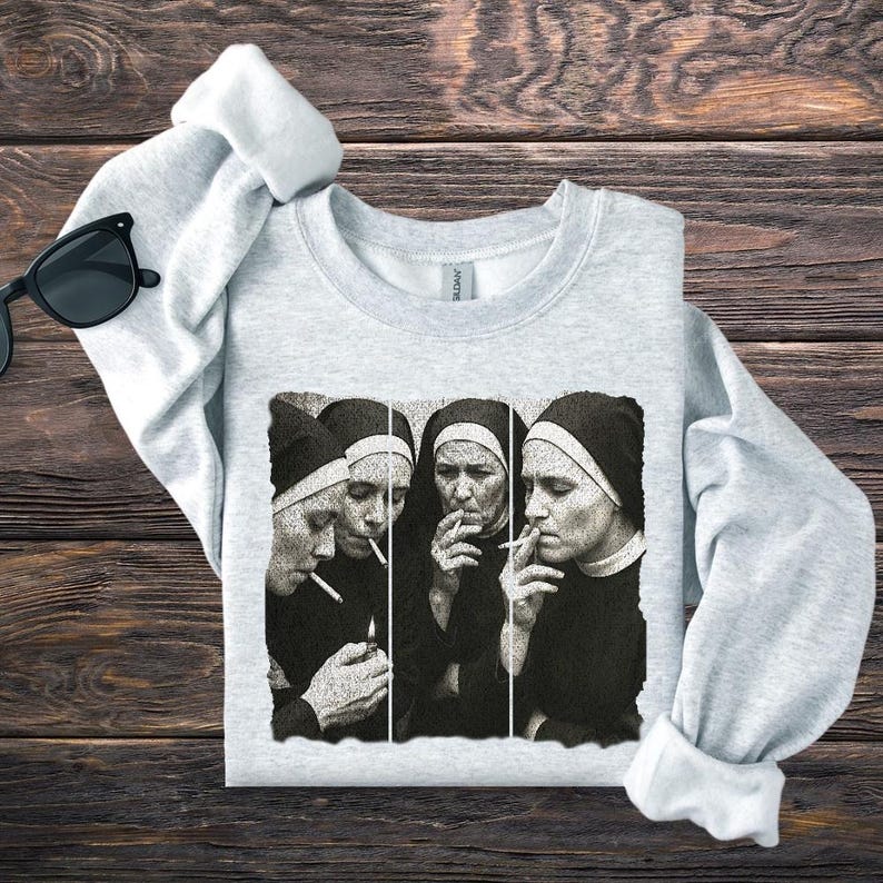 May include: Light gray sweatshirt featuring a black and white graphic of four nuns smoking cigarettes. The image is a stylized, artistic representation. The sweatshirt is folded on a wooden surface, with a pair of black sunglasses visible.