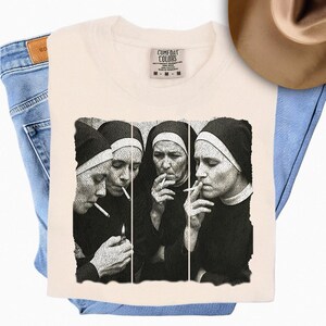 May include: Cream-colored t-shirt featuring a black and white image of four nuns smoking cigarettes. The shirt is folded on top of blue jeans. The Comfort Colors tag is visible at the top of the shirt.