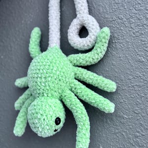 May include: A handmade, plush spider toy in a vibrant green color. The spider has eight long, curved legs and a round body. It features a white, looped hanging cord. The spider's eyes are small, black beads, and it has a simple, smiling face.