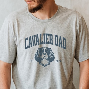May include: A gray t-shirt with the text "Cavalier Dad" in blue, featuring a Cavalier King Charles Spaniel dog illustration and the text "EST 2024".