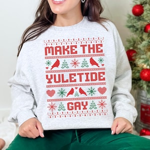 May include: A light gray sweatshirt with the text "MAKE THE YULETIDE GAY" in red. The design includes red birds, snowflakes, and hearts. The person is wearing green pants and is near a Christmas tree.