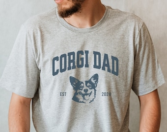 May include: A gray t-shirt with the text "Corgi Dad" in blue lettering. The text is arched over a blue image of a corgi dog. The text "EST 2024" is below the dog image.
