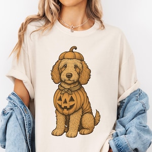 May include: Cream-colored t-shirt featuring a cartoon illustration of a dog dressed as a pumpkin for Halloween. The dog is brown with a pumpkin costume and hat. The design is centered on the shirt, offering a festive and seasonal theme.