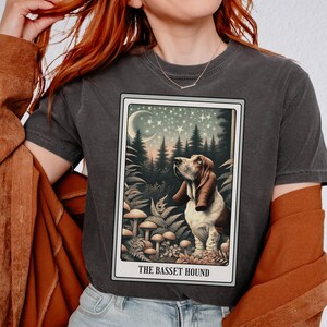 May include: Dark gray t-shirt featuring a tarot card design with a Basset Hound illustration. The card depicts a forest scene with mushrooms, ferns, and a starry night sky. The text "THE BASSET HOUND" is at the bottom of the card.