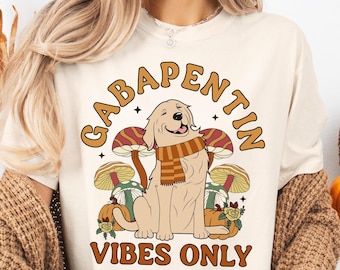 Könnte beinhalten: Cremefarbenes T-Shirt mit dem Text "GABAPENTIN VIBES ONLY" in Braun. Die Grafik zeigt einen lächelnden Golden Retriever mit Schal, Pilzen und Kürbissen. Das Design hat eine Retro-Ästhetik zum Thema Herbst.