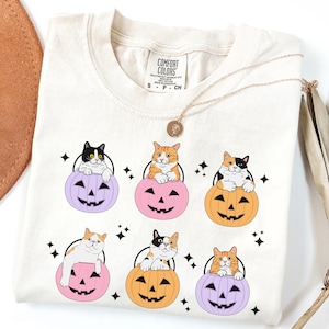 May include: Cream-colored t-shirt featuring six cartoon cats perched atop jack-o'-lanterns in shades of orange, pink, and purple. The cats have various markings, and the shirt has a small tag that says "Comfort Colors". A gold necklace is draped across the shirt.