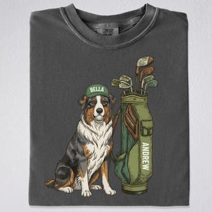 May include: Dark gray t-shirt featuring a cartoon illustration of a dog wearing a green cap with the name "Bella" and a golf bag with the name "Andrew". The dog is sitting next to the golf bag filled with golf clubs.