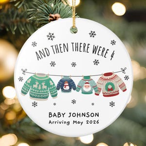 May include: A white ceramic Christmas ornament with the text "AND THEN THERE WERE 4" and "BABY JOHNSON Arriving May 2026". The ornament features four sweaters hanging on a clothesline, with snowflake accents.
