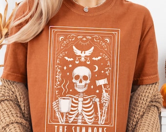 May include: A rust-colored short-sleeve t-shirt featuring a white graphic print of a skeleton holding a coffee mug and a gavel. The design is framed like a tarot card with celestial symbols, bats, and the text THE SUMMONS at the bottom.