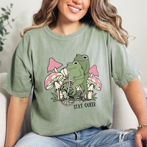 May include: A green t-shirt with a graphic of two frogs riding a bicycle with mushrooms in the background. The text "STAY QUEER" is printed below the frogs.