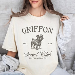 May include: A beige t-shirt with a black graphic of a pug dog. The graphic says "Griffon Social Club" with the text "Est. 2024" and "San Francisco, CA".