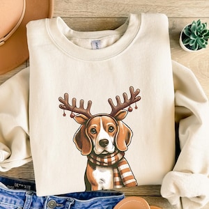 May include: A beige sweatshirt with a cartoon image of a beagle dog wearing a red and brown reindeer antler headband with red berries. The dog is wearing a striped scarf and has a brown nose.