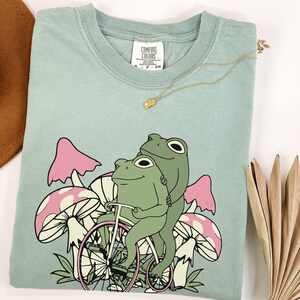 May include: A light blue t-shirt with a graphic of two green frogs riding a bicycle through a field of pink and white mushrooms. The shirt has the text "Comfort Colors" printed on the tag.