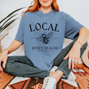 May include: A blue t-shirt with a black graphic design that says "LOCAL" with a bee illustration and the text "EST. 2024 HONEY DEALER SAN FRANCISCO, CA".