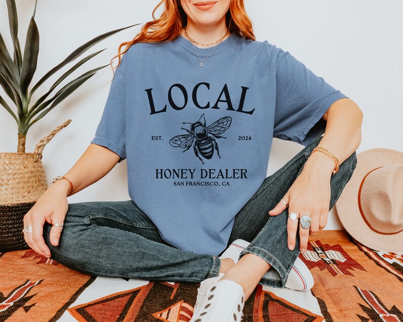 May include: A blue t-shirt with a black graphic design that says "LOCAL" with a bee illustration and the text "EST. 2024 HONEY DEALER SAN FRANCISCO, CA".