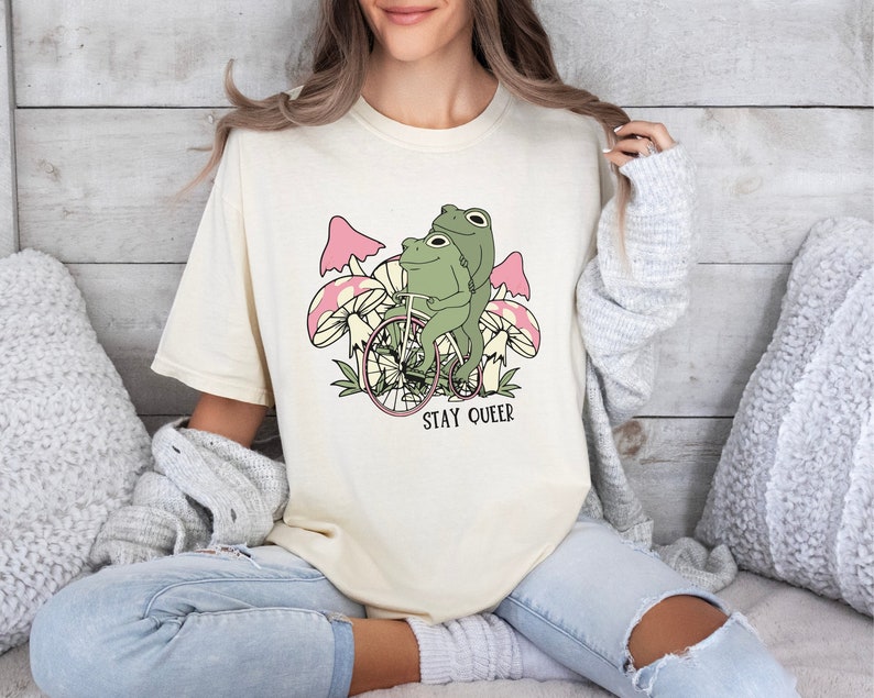May include: A cream colored t-shirt with a graphic of two green frogs riding a bicycle through a field of pink mushrooms. The text "STAY QUEER" is printed below the image.