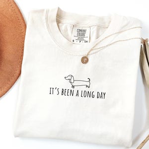 May include: Cream-coloured t-shirt featuring a black dachshund illustration and the text "It's Been a Long Day." A gold necklace with a pendant is on the shirt. The shirt has a Comfort Colors tag.