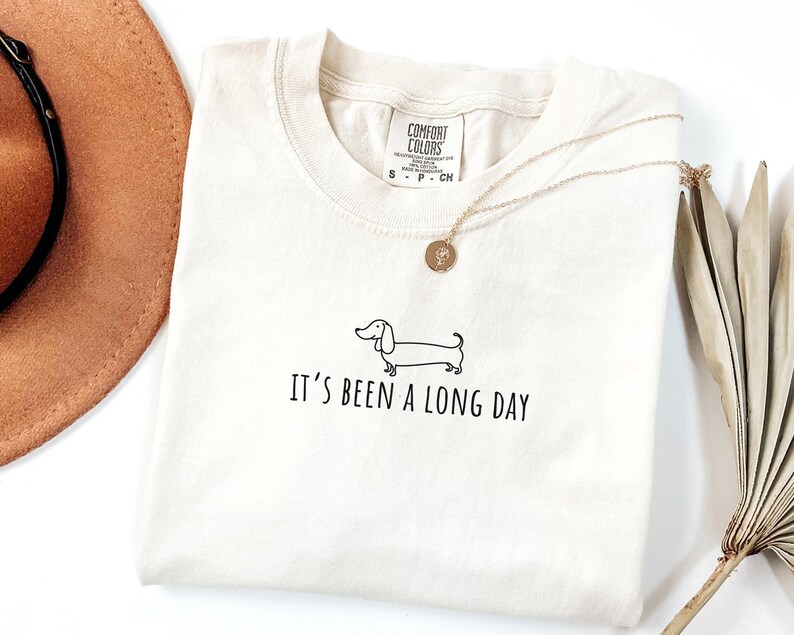 May include: Cream-coloured t-shirt featuring a black dachshund illustration and the text "It's Been a Long Day." A gold necklace with a pendant is on the shirt. The shirt has a Comfort Colors tag.