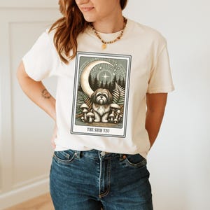 May include: A white t-shirt with a graphic of a Shih Tzu dog in a forest setting with a crescent moon and stars. The text "THE SHIH TZU" is printed below the image.