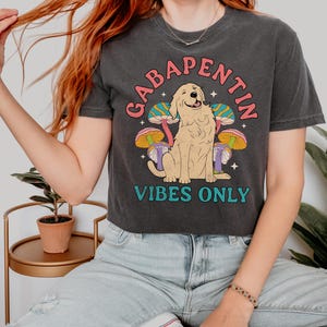 K&ouml;nnte beinhalten: Ein schwarzes, bauchfreies T-Shirt mit einem Aufdruck eines Golden Retrievers, der vor Pilzen sitzt. Der Text "Gabapentin Vibes Only" ist auf dem Shirt gedruckt.