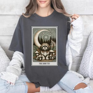 May include: A dark gray t-shirt with a graphic of a Shih Tzu dog sitting in a forest with mushrooms and a crescent moon. The text "THE SHIH TZU" is printed below the graphic.